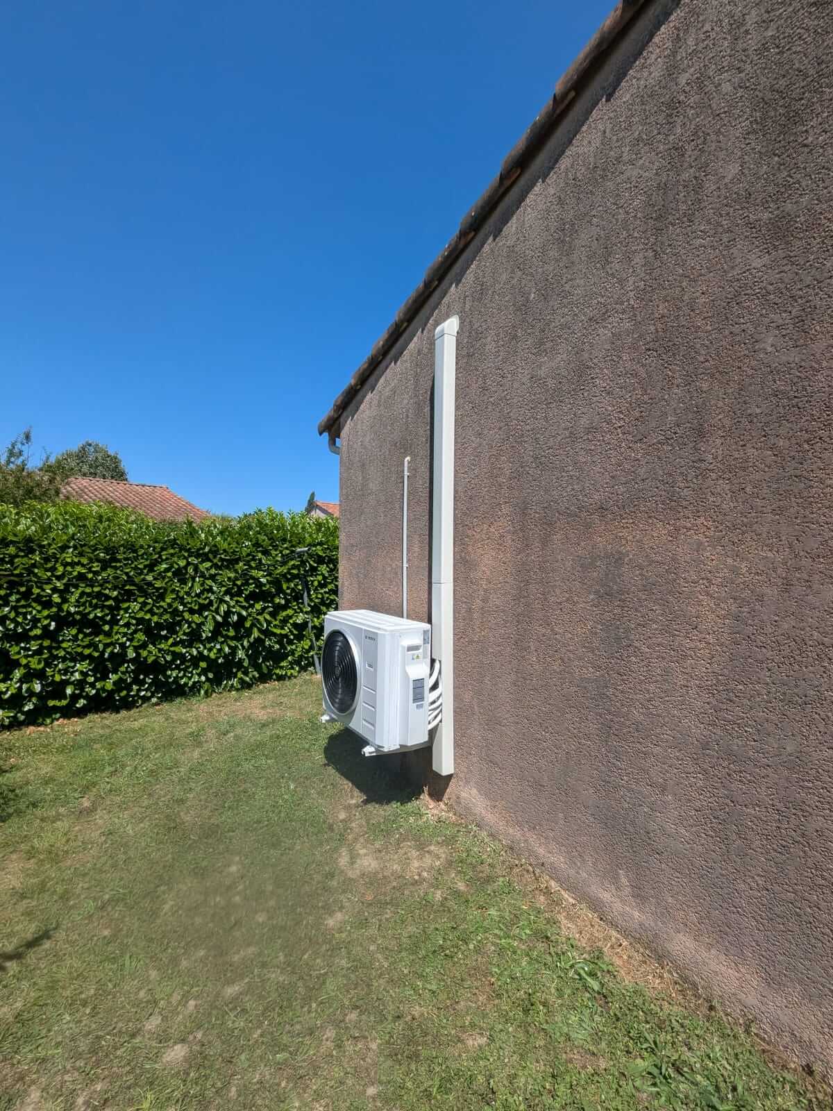 Une clim réversible Bosch quadri-split blanc est fixé sur le mur extérieur d’une maison en crépi marron. L’appareil est placé à environ cinquante centimètres du sol, relié par des gaines blanches qui montent le long du mur. Au sol, il y a de l’herbe, et en arrière-plan on distingue une haie verte ainsi que le toit d’une autre maison. Le ciel est bleu et dégagé.