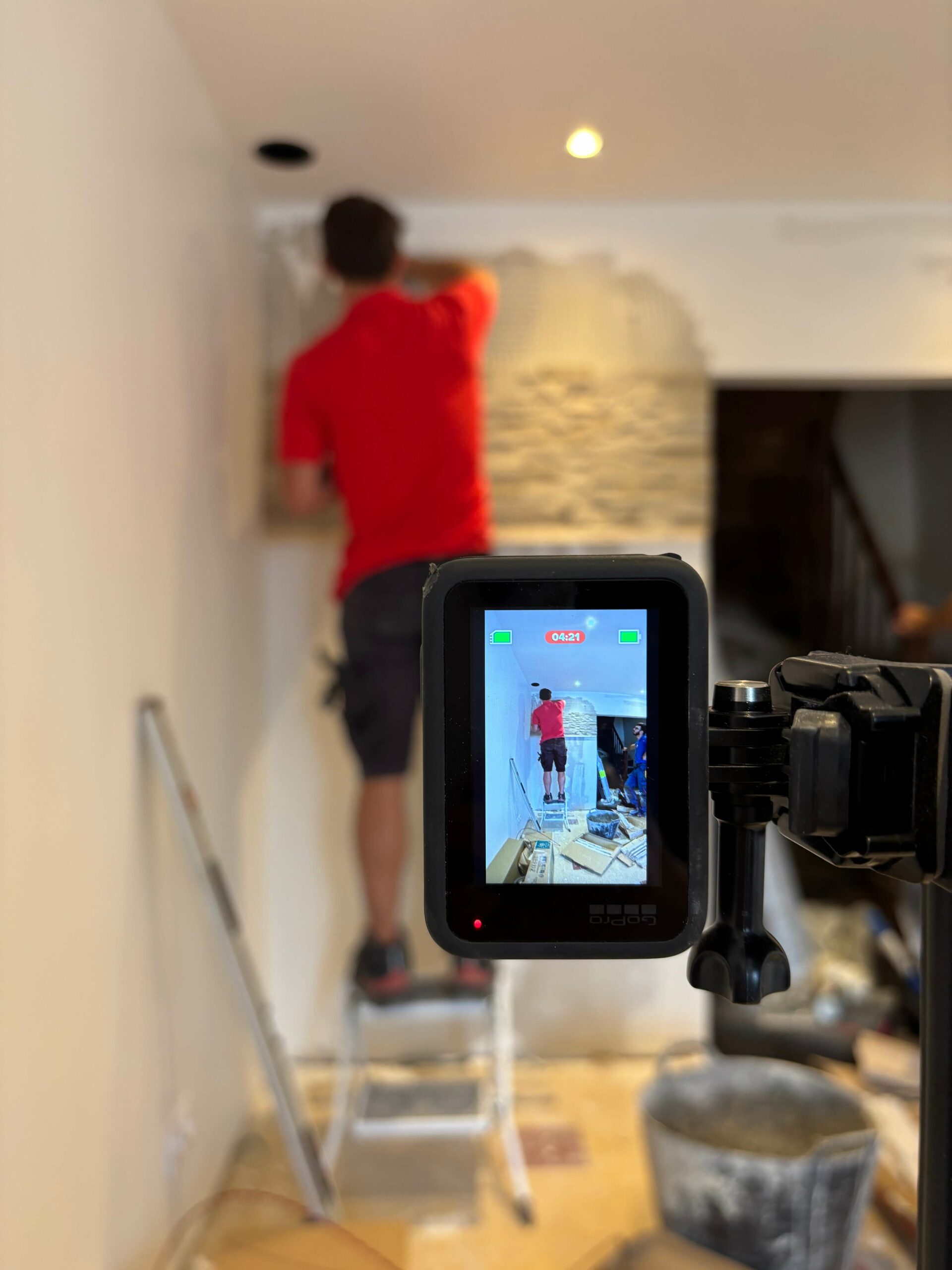 Dans une pièce en rénovation, un homme en tee-shirt rouge et short noir se tient sur un escabeau et applique de la colle sur un mur. Le mur est en partie recouvert de colle et de pierres décoratives claires déjà posées. Au sol se trouvent un seau et du matériel de chantier. Au premier plan, une caméra filme la scène et affiche l’homme en train de travailler sur le mur.