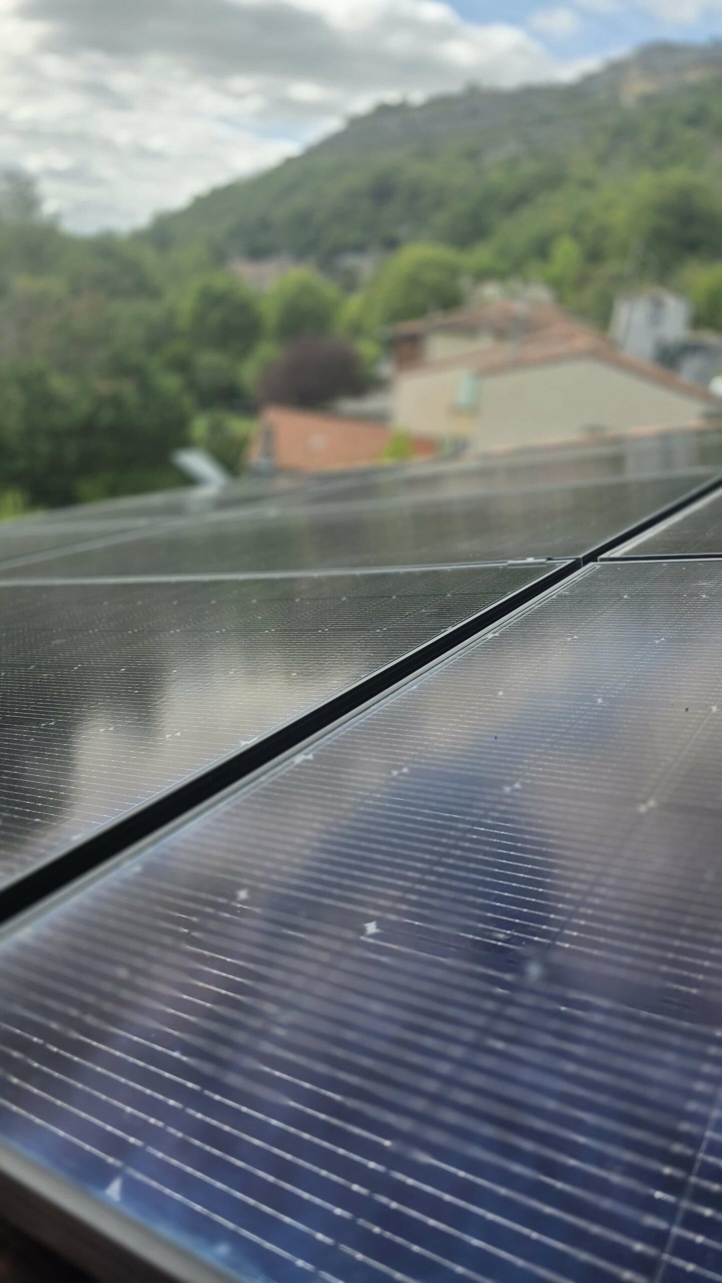 Les panneaux solaire occupent la majorité de l’image. Ils sont rectangulaires, noirs, avec de fines lignes parallèles blanches qui traversent leur surface. Ils sont légèrement inclinés, ce qui laisse penser qu’ils sont installés sur un toit. L’arrière-plan est flou, derrière les panneaux, on distingue un paysage un peu flou à cause de la profondeur de champ de l’appareil photo. On devine : - Des maisons aux toits orangés et aux murs clairs. - Beaucoup de végétation verte, des arbres touffus qui couvrent une grande partie du décor. - Une colline boisée qui monte vers le ciel, donnant une impression de village entouré par la nature. - Le ciel est partiellement nuageux, laissant apparaître des zones claires et lumineuses mais aussi des nuages gris-blanc.